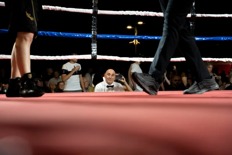 Inquadratura dal bordo del ring: arbitro in frac e pubblico sullo sfondo durante la serata di pugilato