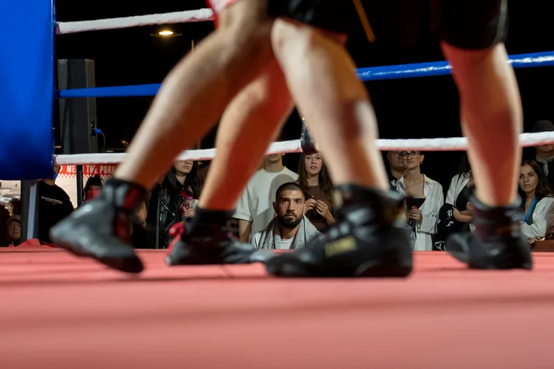 L'istruttore segue concentrato l'incontro dal bordo del ring, incorniciato dai piedi dei pugili in primo piano